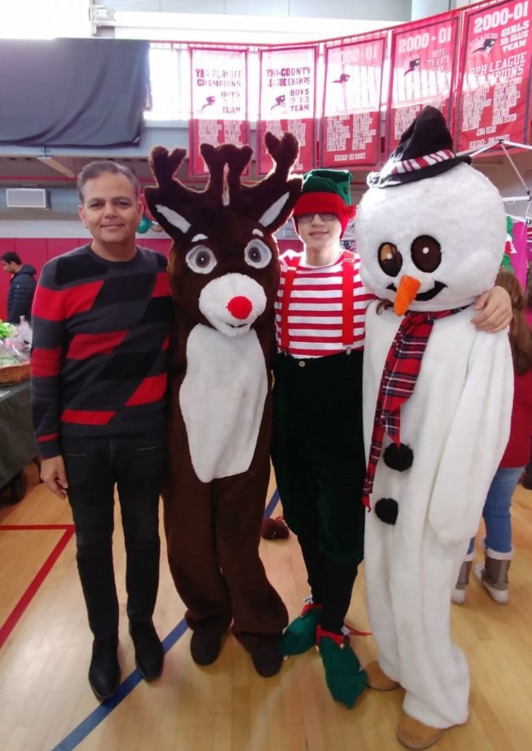 Raj at a holiday community event with reindeer, elf and snowman mascots in the school gym