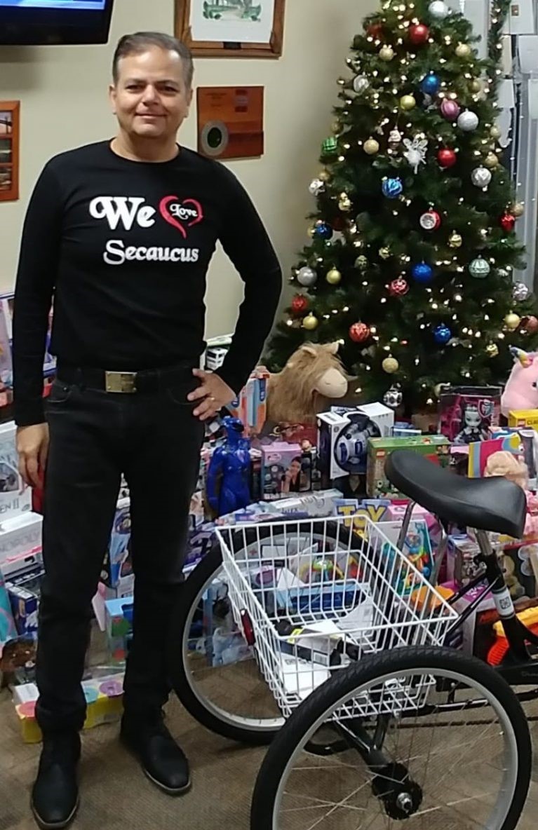 Raj in a 'We Love Secaucus' shirt standing beside a mountain of donated toys and a new tricycle
